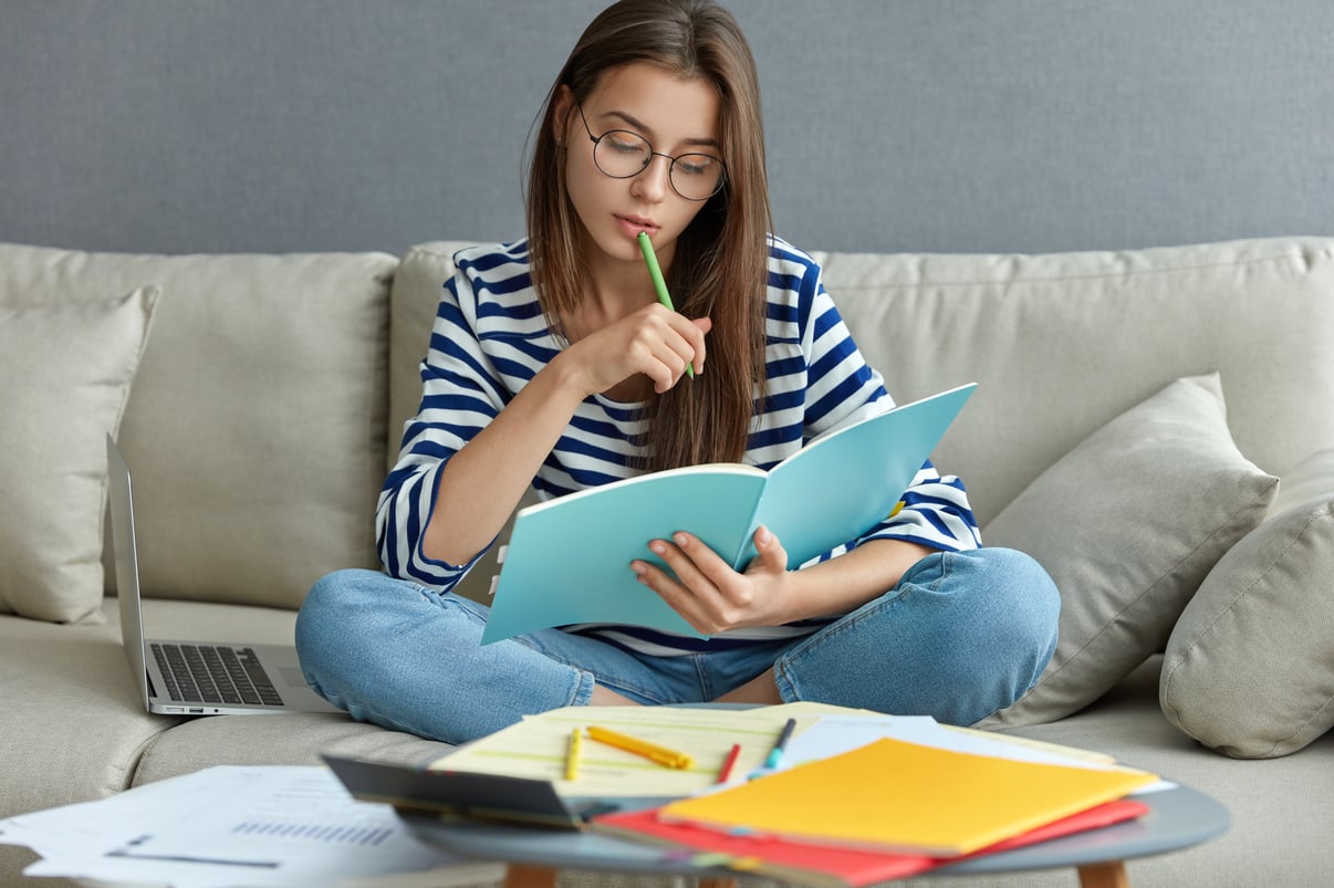 estudiar-concepto-linea-mujer-joven-seria-que-ocupada-proyecto-independiente-remoto-sienta-comodo-sofa-escribe-notas-sostiene-libro-texto-usa-computadora-portatil-casa-internet-inalambrico